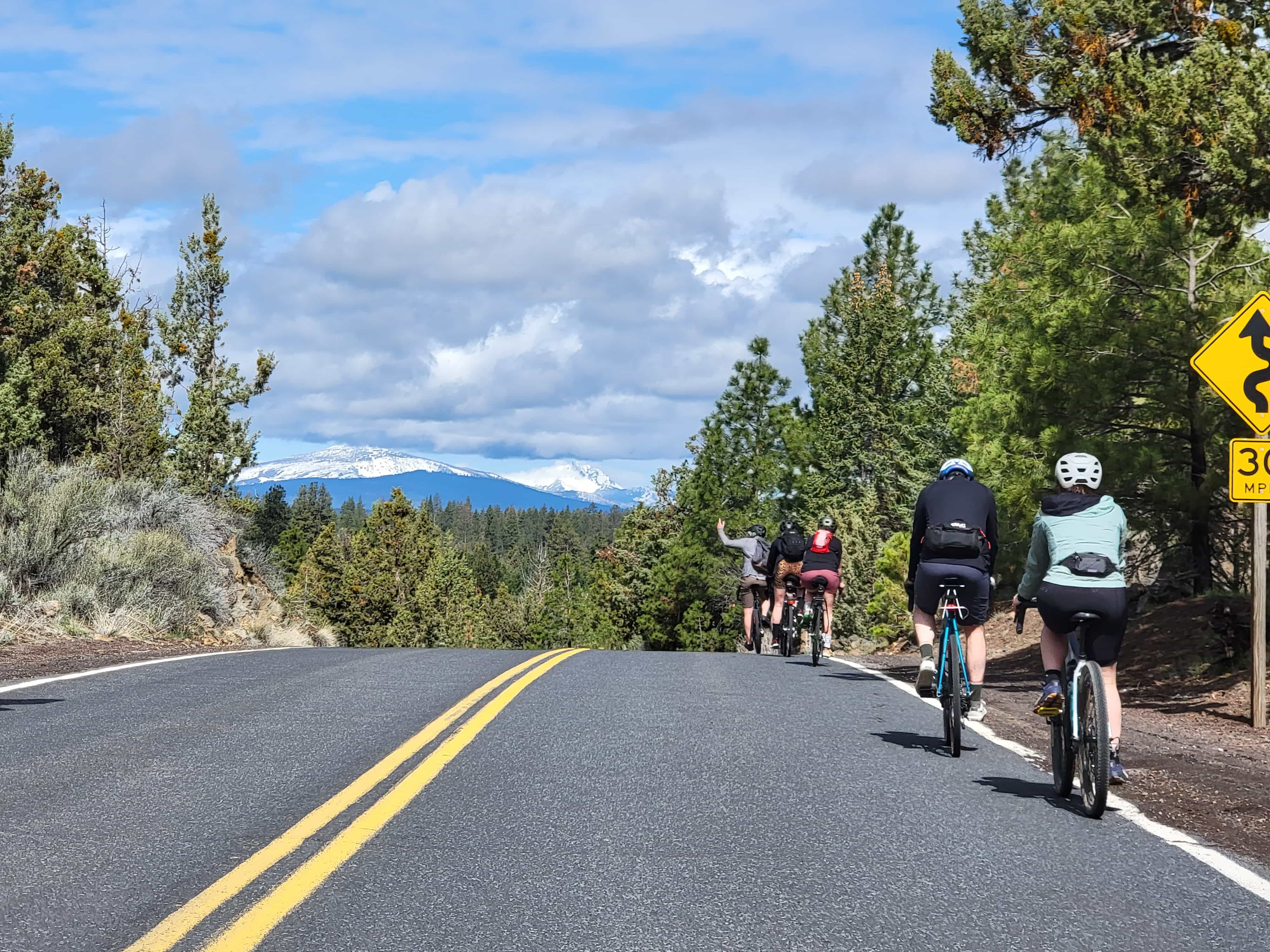 A picture taken from behind of 4 ciclists biking on a back road, about to head downhill, blue skies and green trees loom overhead